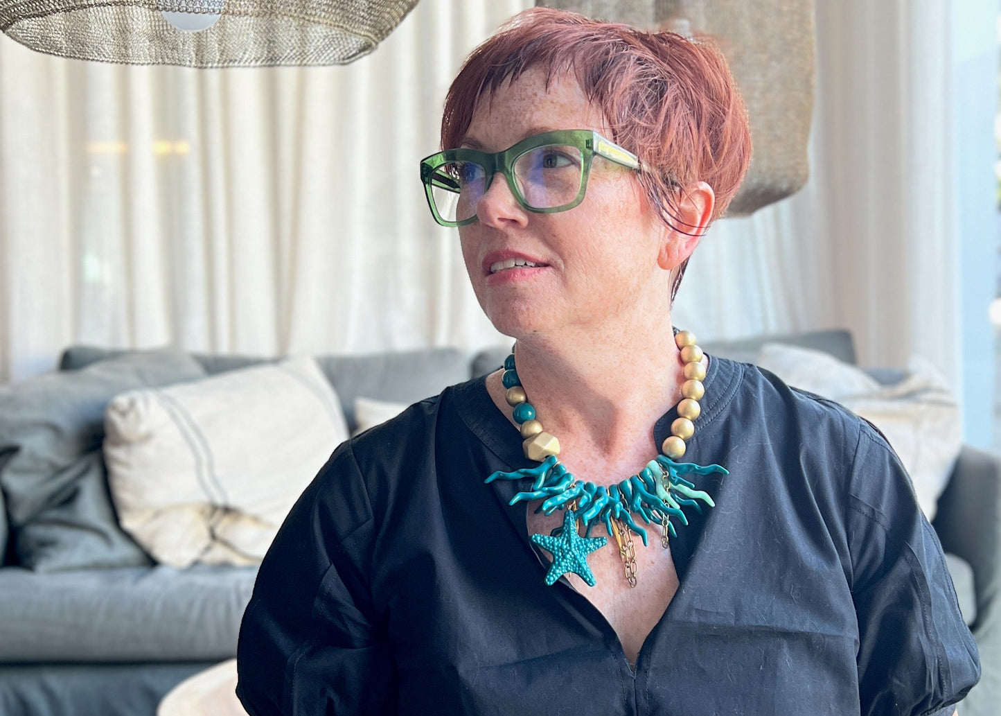 Over 50 modern woman with red hair and glasses sitting in a modern living room with edgy coastal chic necklace.